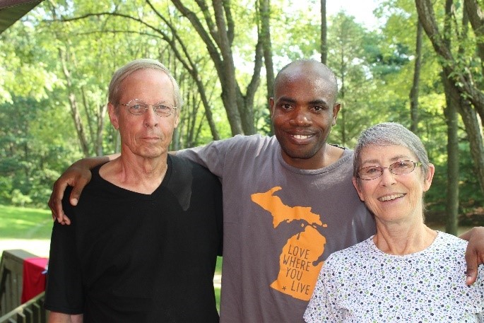 Three people standing together, the person in the center is Juma and he has his hands around their shoulders. Green foilage is in the background.