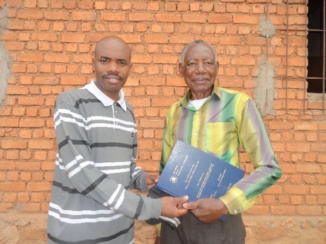 A parent and adult child stand together holding each other's hands. The parent is also holding the son's doctorial thesis.