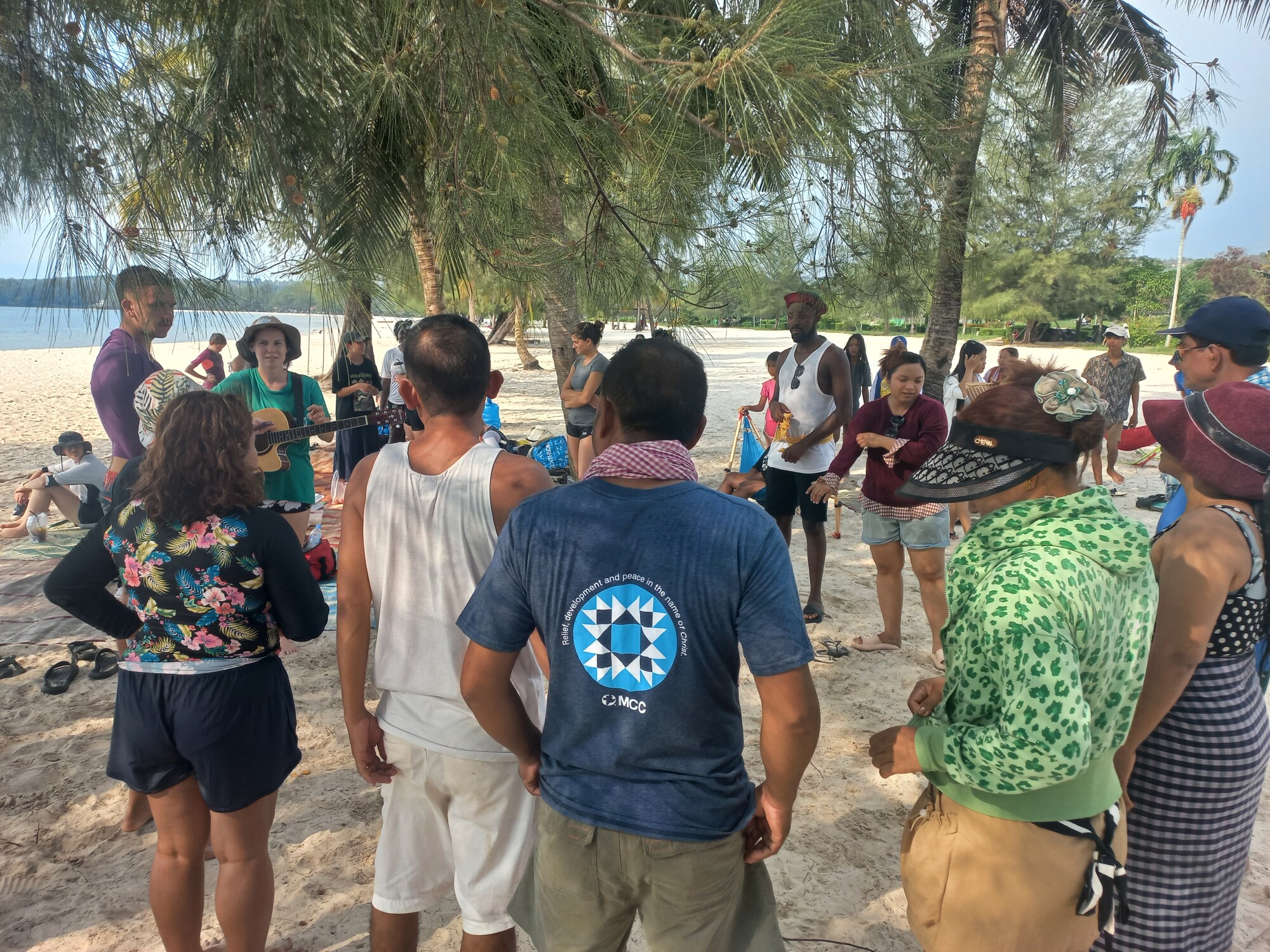 A group of people is gathered on a beach with trees, some facing away from the camera. They appear to be engaged in a social activity or event, with a clear sky above.