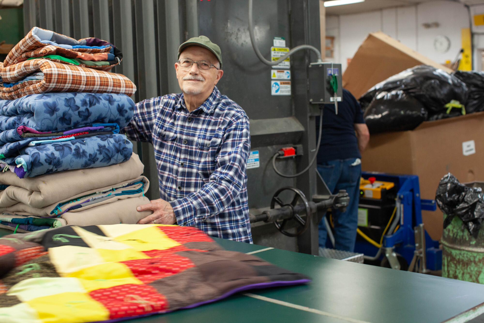 An elderly man in a cap and plaid shirt stands beside a stack of folded blankets in an industrial setting, possibly a warehouse or factory.