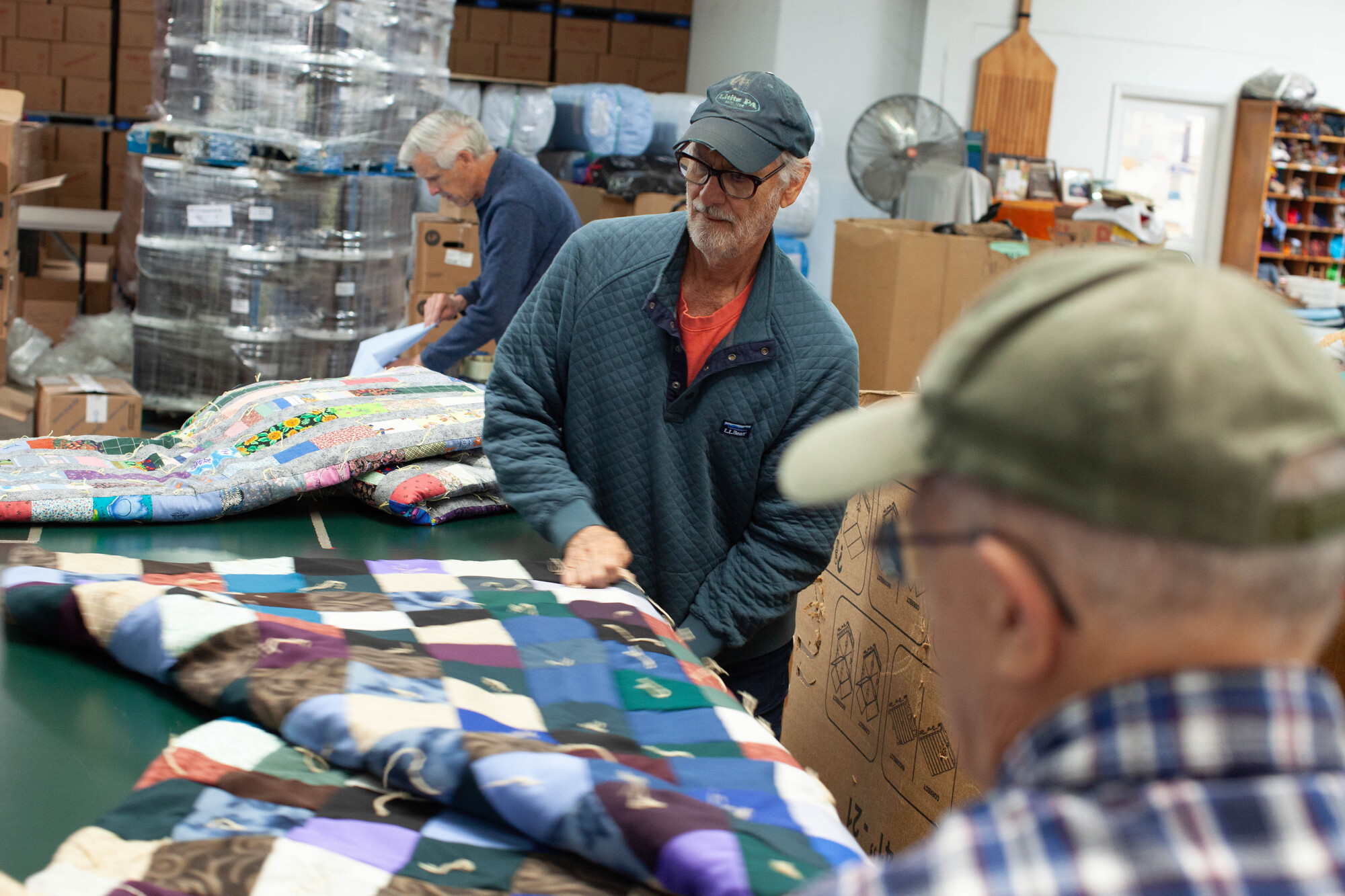 Men in a workshop inspect a quilt. One in focus wearing glasses and a cap, others in the background, with boxes and industrial setting.