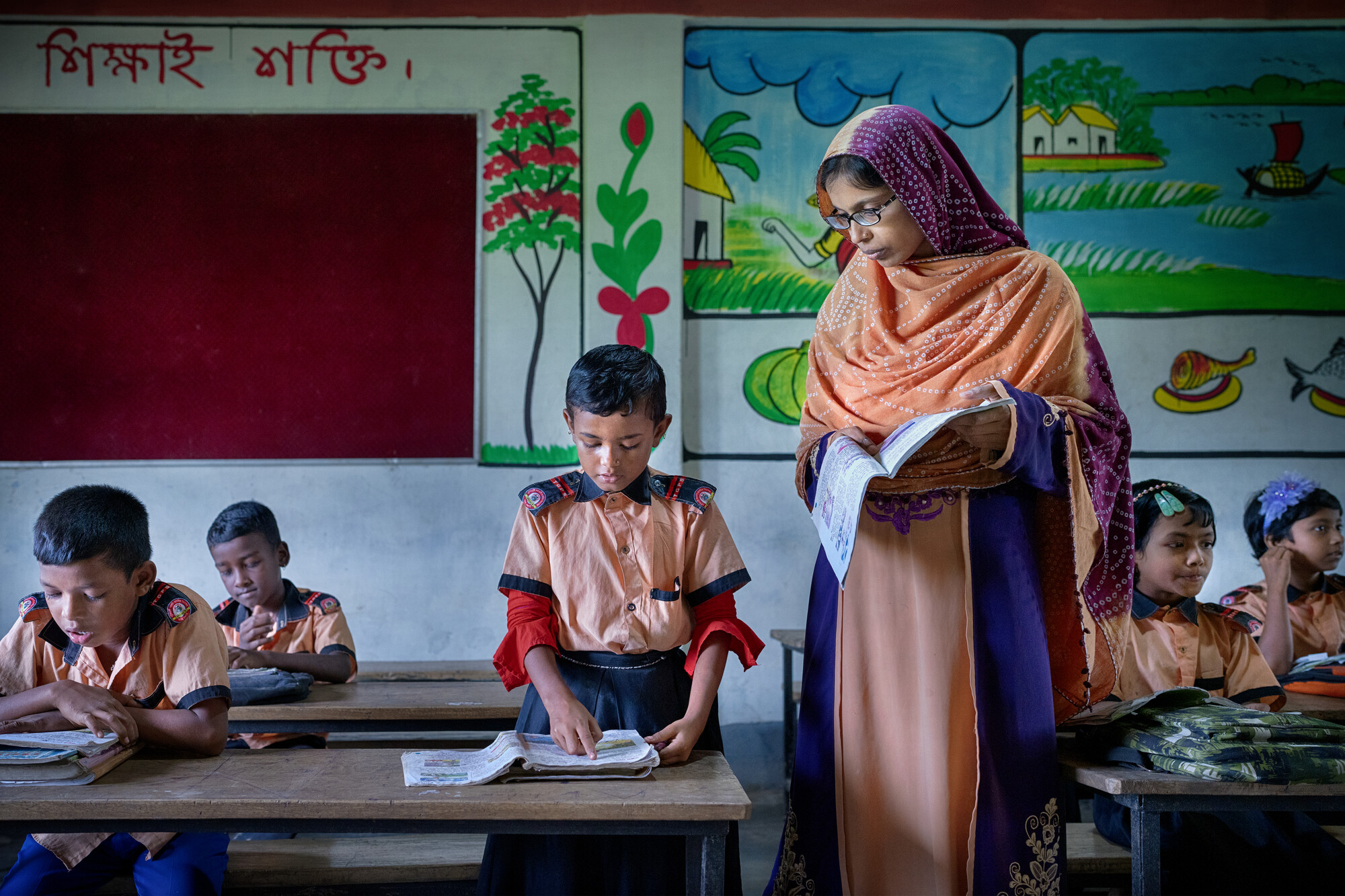 A student stands and recites from a book while a teacher watches him.