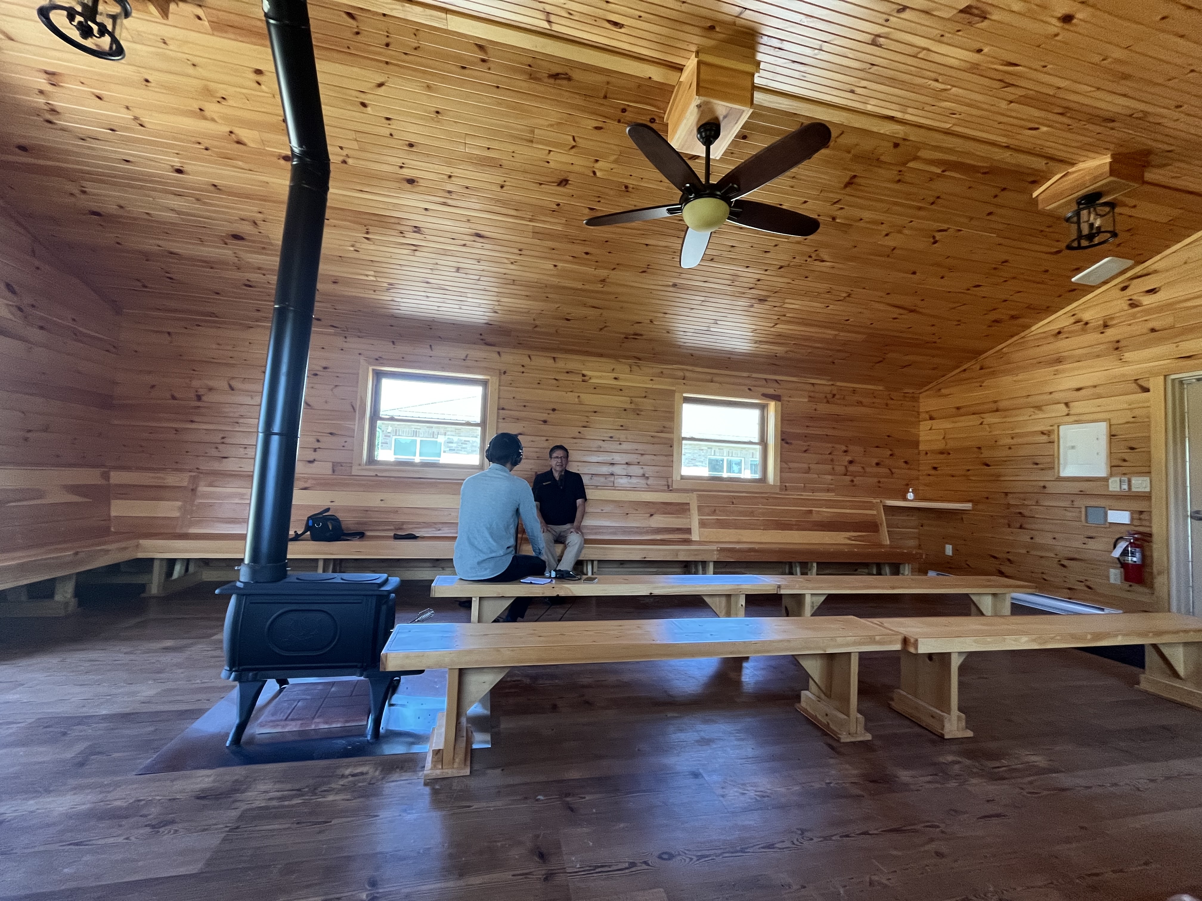 A man in green shirt interviews a man in black shirt in a one-room building clad in wood with a black wood stove in foreground