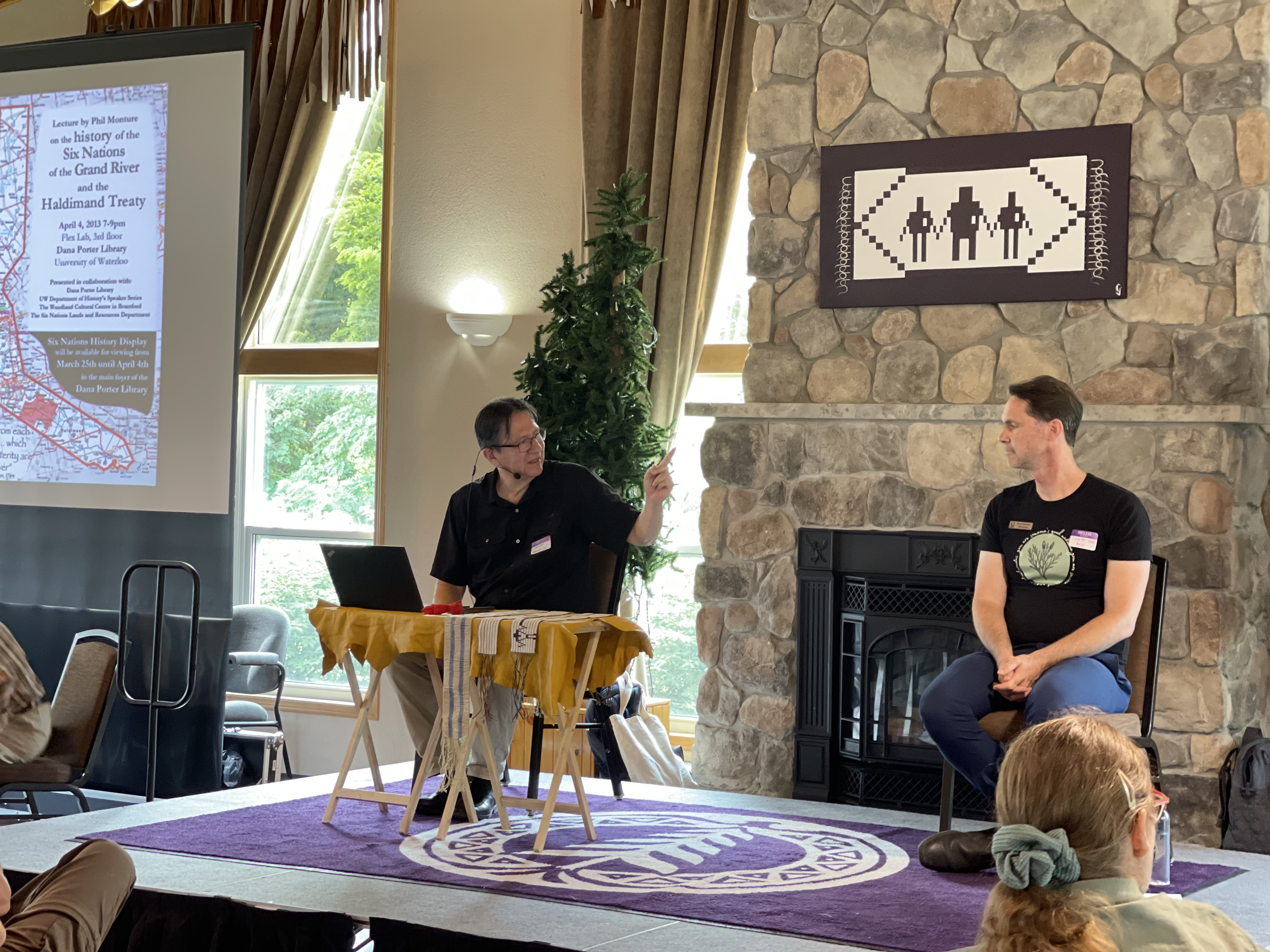 Two men on a stage, one on the left with wampum belts on the table in front of him