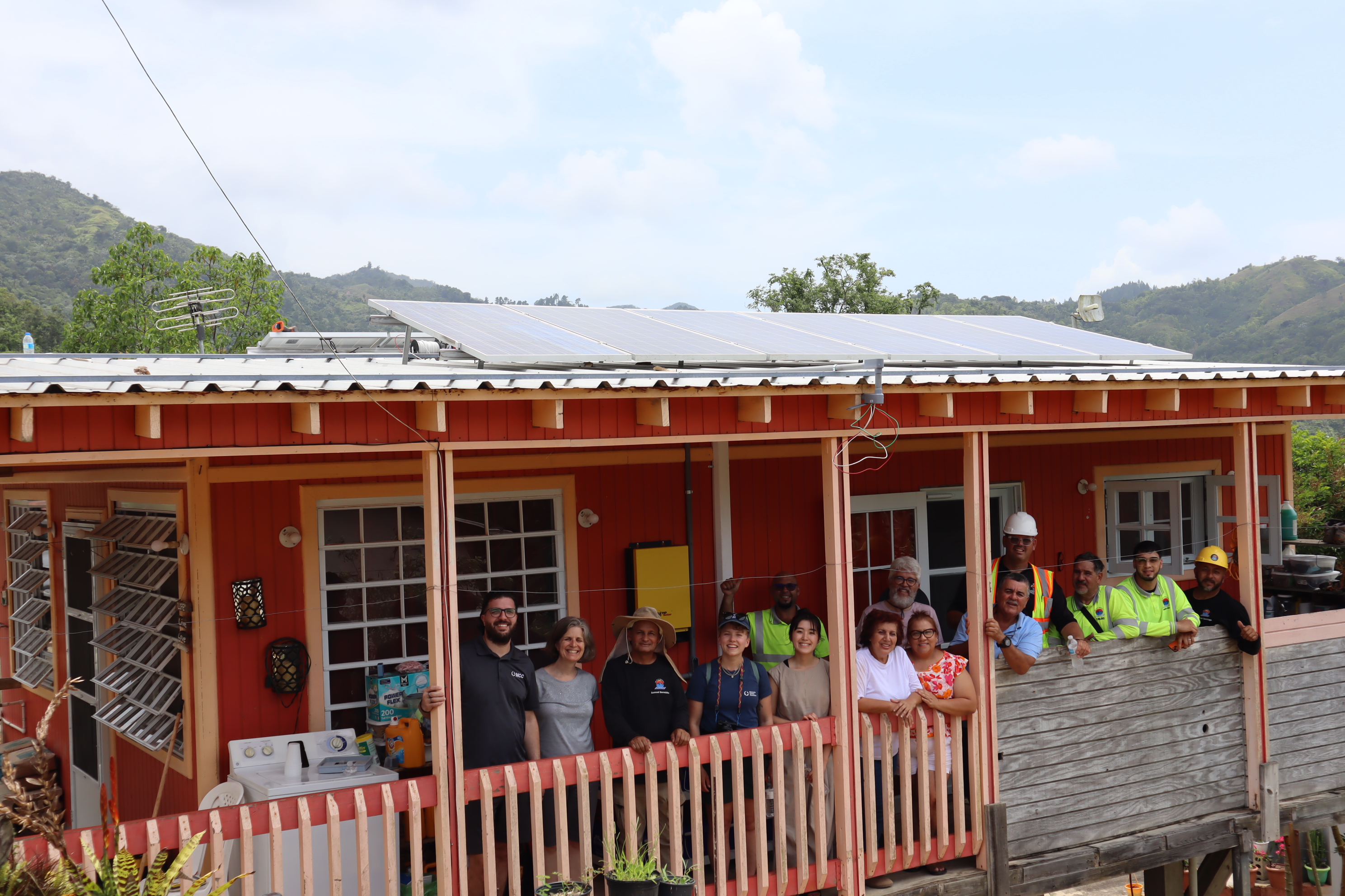 a group of MCC staff and partners gather on the porch of a house where solar panels were just installed