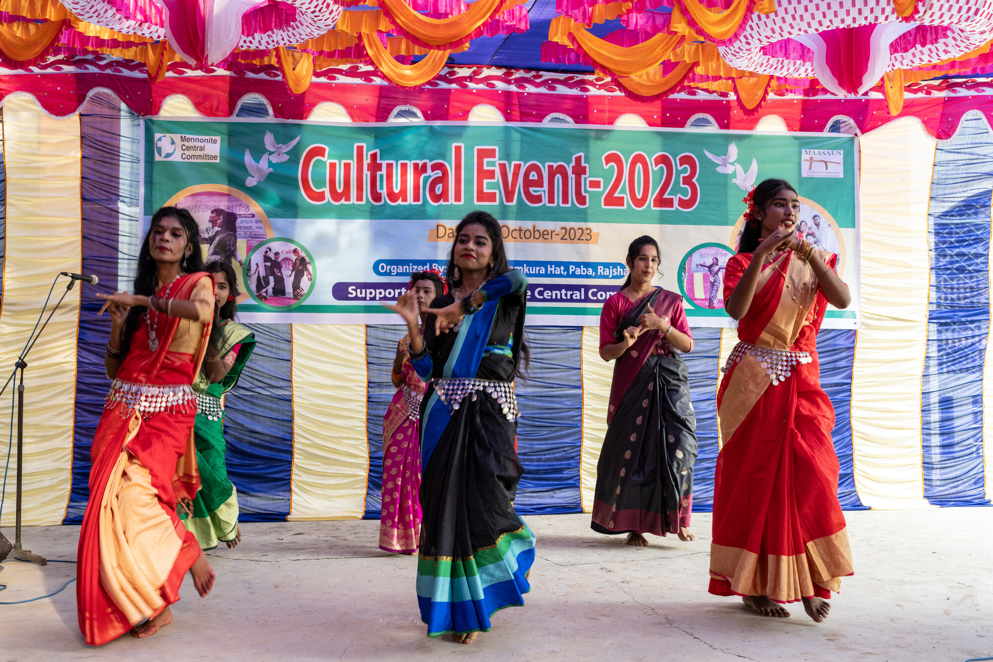 Four dancers in colorful traditional attire perform at a "Cultural Event-2023" against a decorated stage backdrop with banners and festoons.