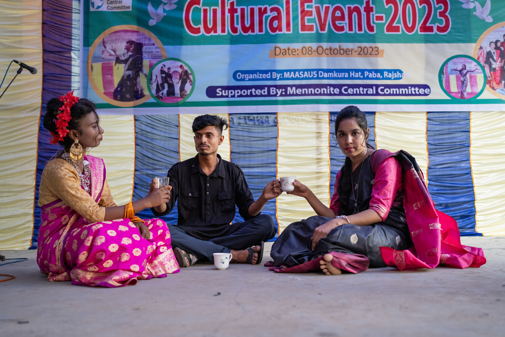 Three individuals are seated on the ground at a cultural event, engaging in an activity. They are dressed in traditional attire, with a banner in the background indicating the event's details.