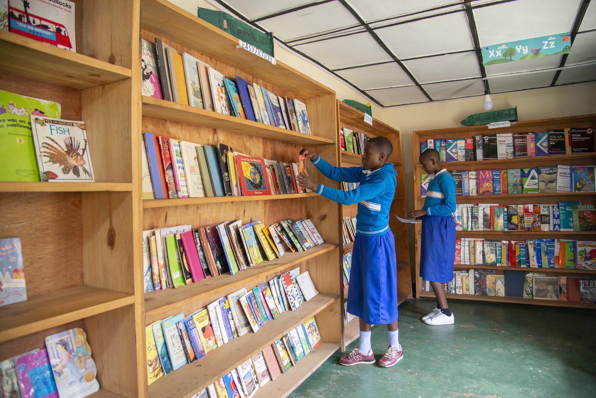 Sonia Uwikunda and Sonia Hakizimana Iraboneye select books at the Gicumbi Children's Peace Library, operated by Transformational Leadership Center. Books are organized into categories like easy fiction, math and science.