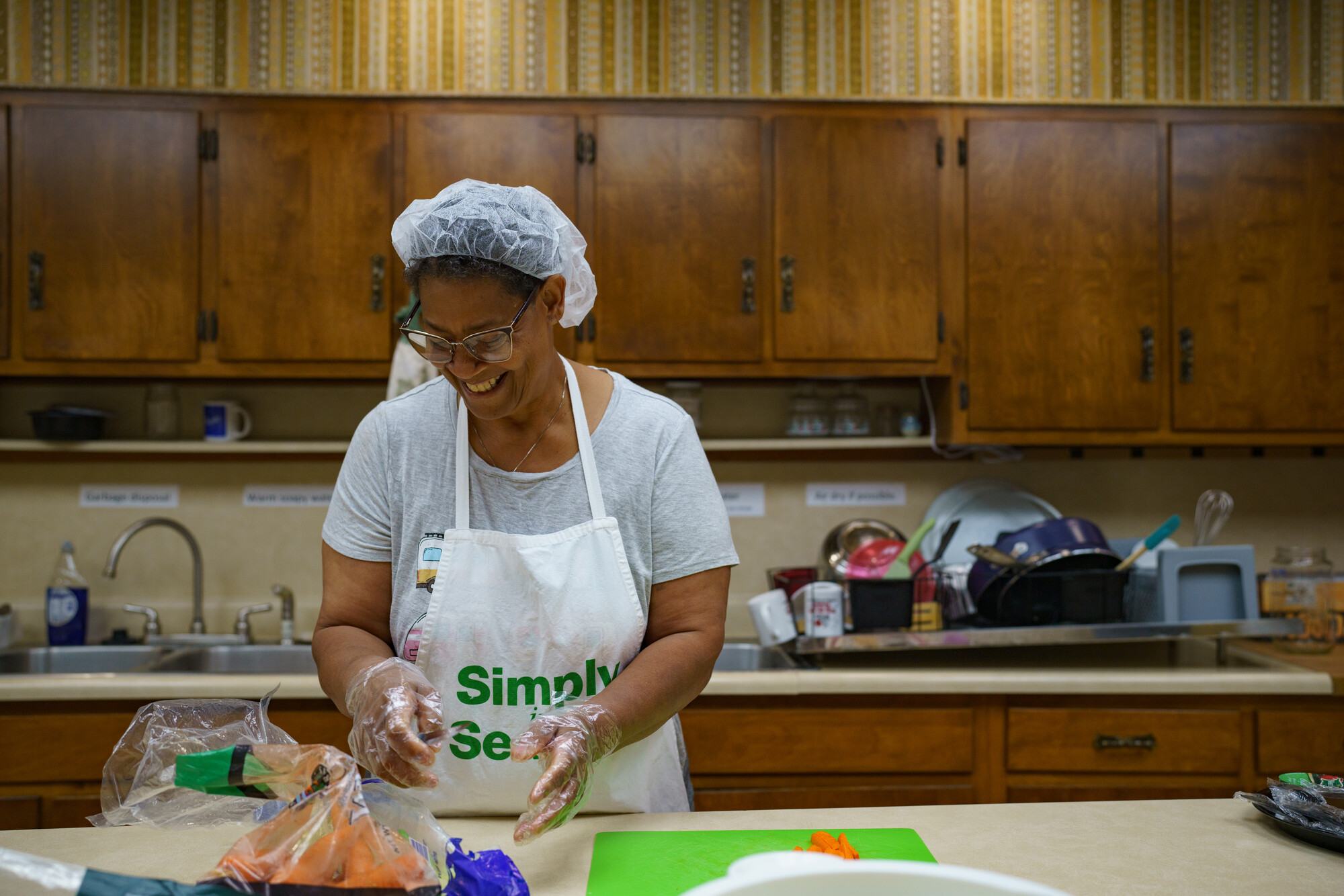 A woman wearing a hairnet and apron is preparing food in a kitchen with wooden cabinets. She is focused on her task, surrounded by cooking utensils and ingredients.