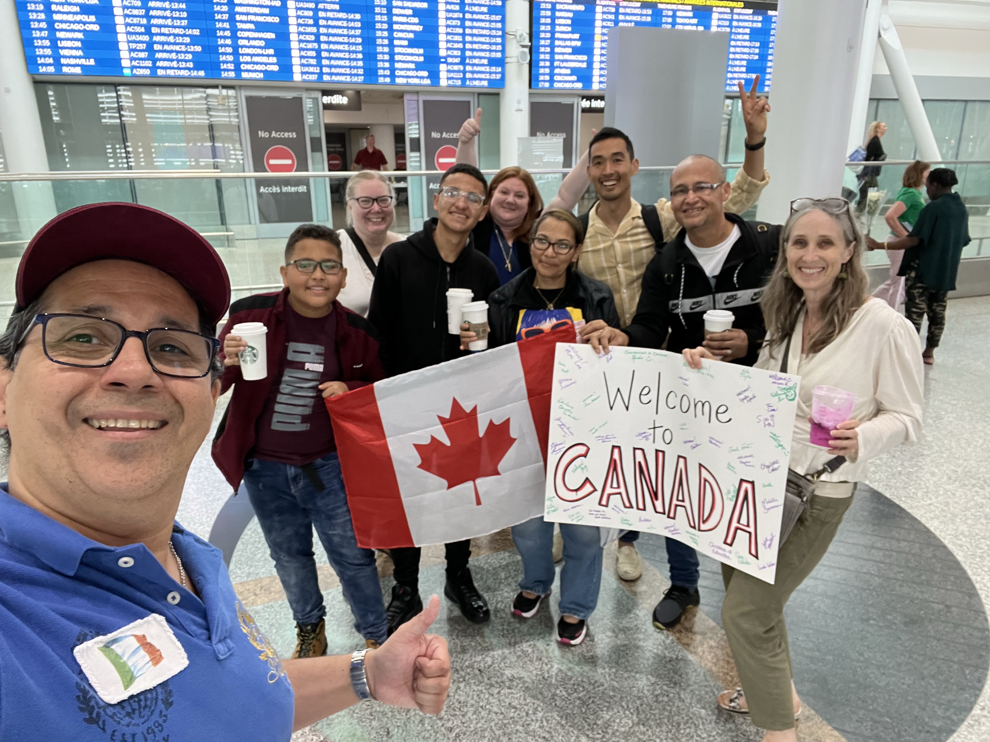 A selfie with smiling group of people at an airport terminal holding signs that read "Bienvenidos" and "Welcome to Canada".