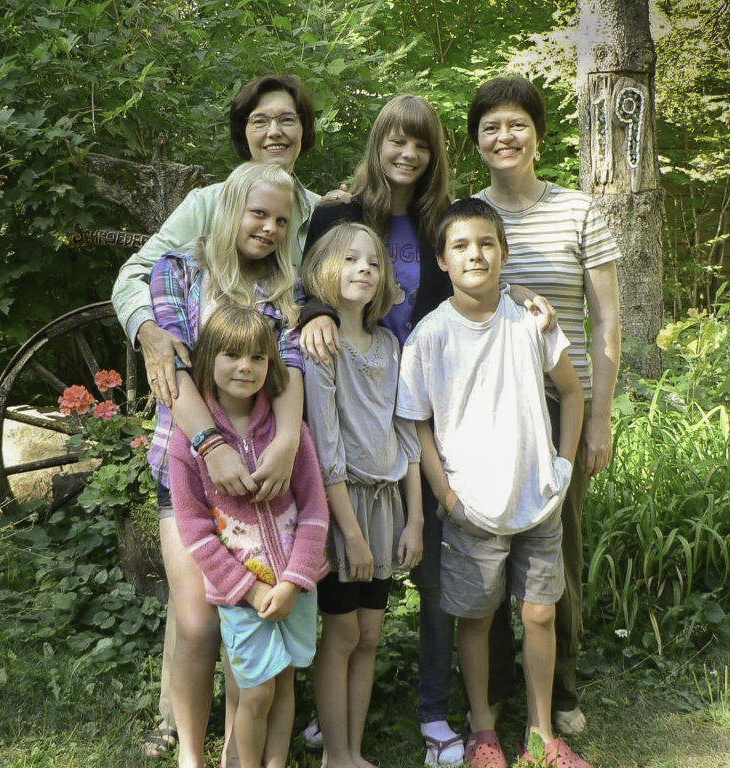 A group of seven individuals, including adults and children, gathered outdoors. They are smiling and standing closely together, surrounded by greenery and plants, with a rustic backdrop that includes an old wheel and decorative elements.