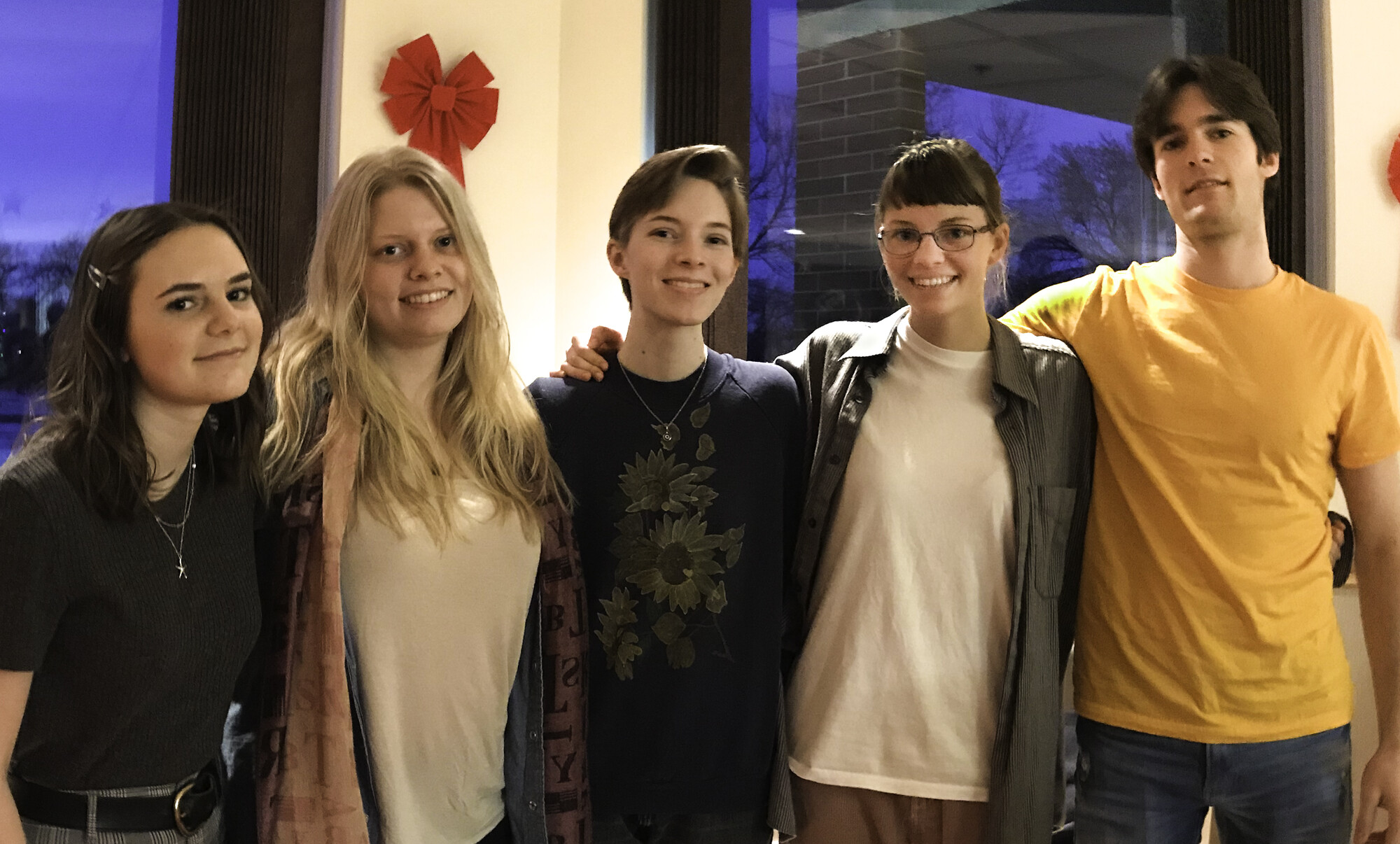 The photograph features five young individuals standing together, smiling and posing in a casual setting, with a festive decoration visible in the background, suggesting a celebratory atmosphere. Their expressions convey warmth and camaraderie.