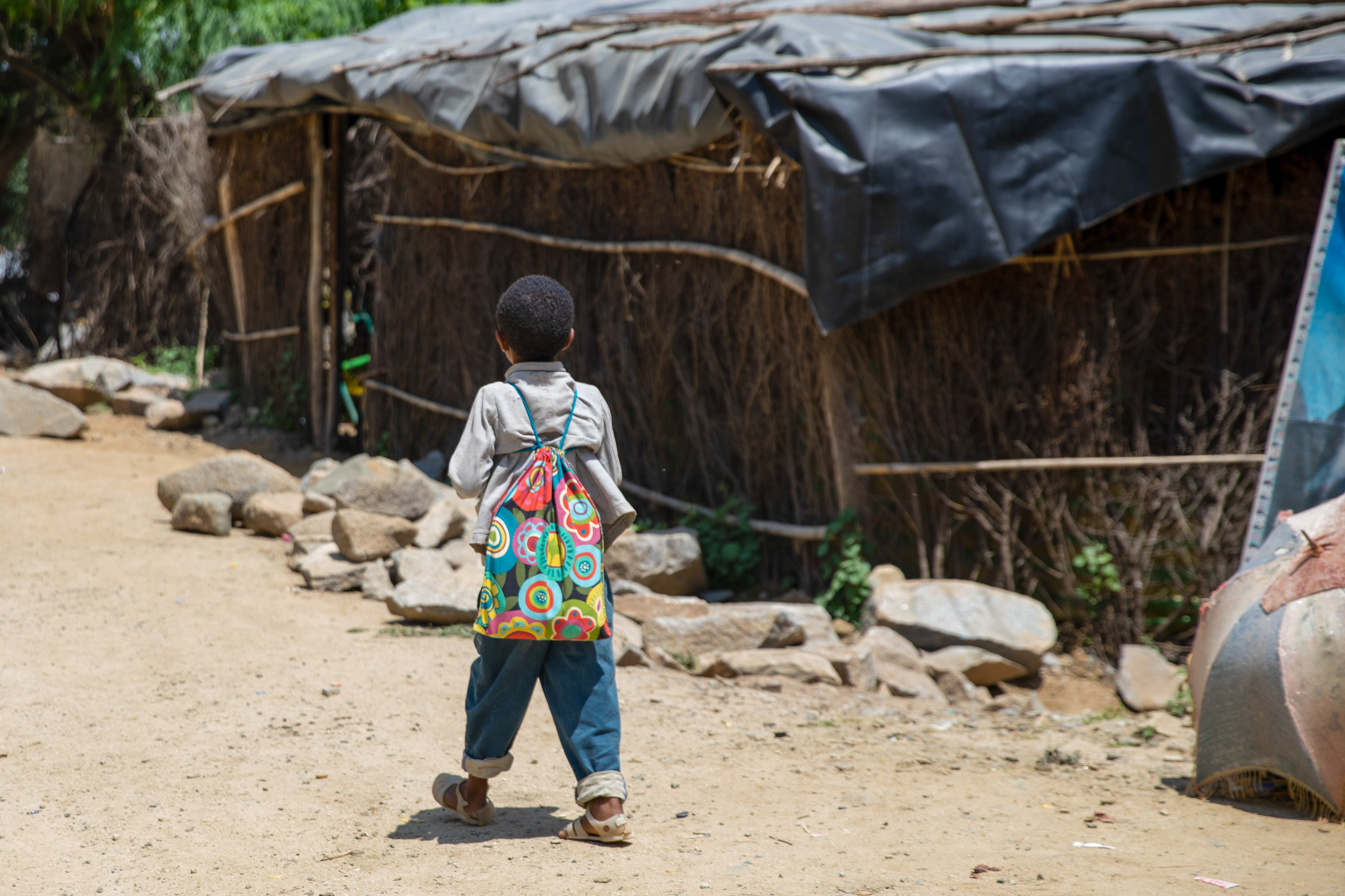 boy carrying school kit like a backpack walking home on dirt roads by stick building