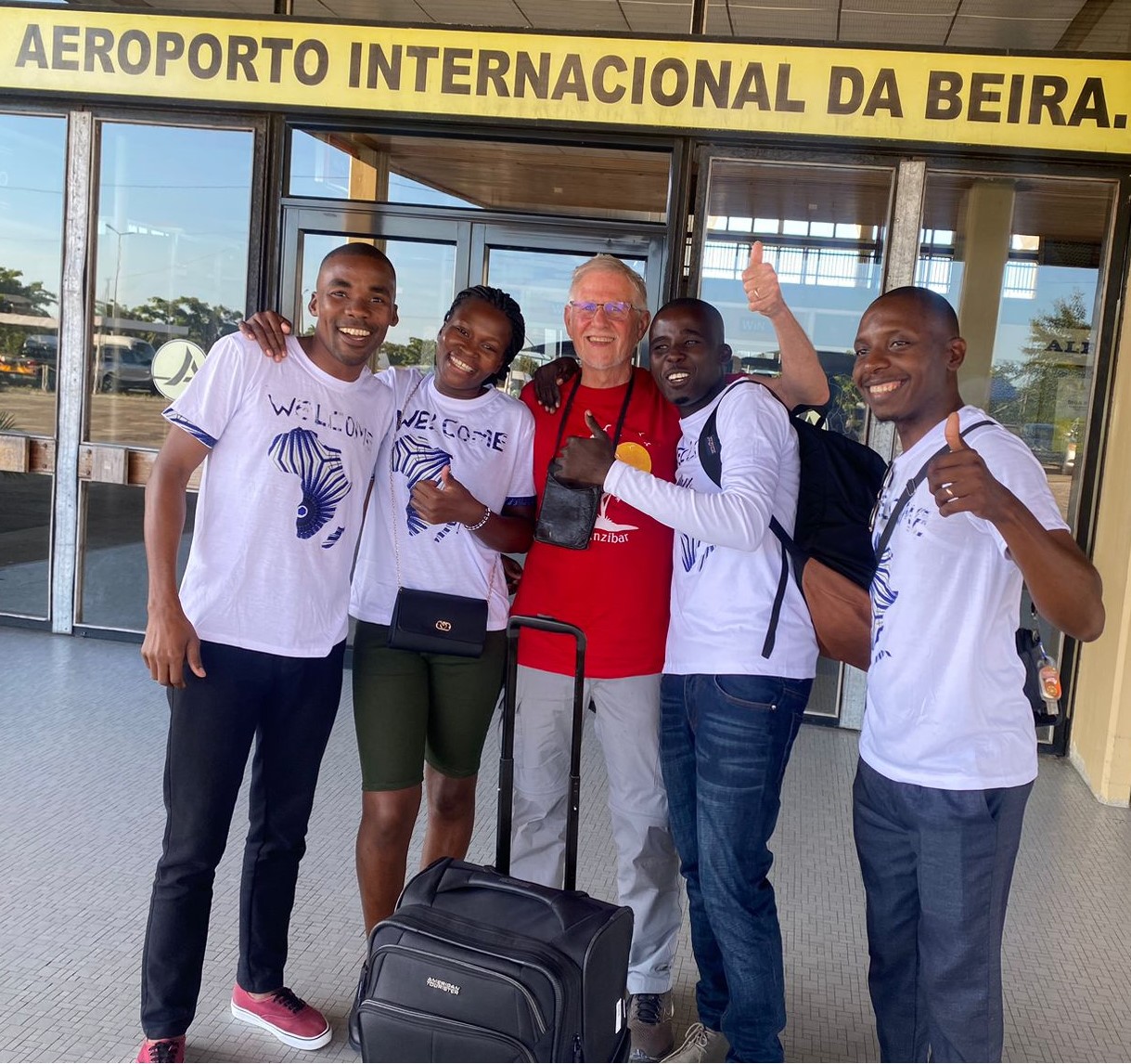 Five people stand outside the Beira International Airport. The four people flanking the person in center are wearing t-shirts that say "welcome."