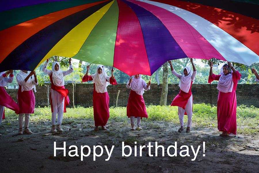 A group of girls lifting a colorful parachute overhead and the words "Happy birthday!"