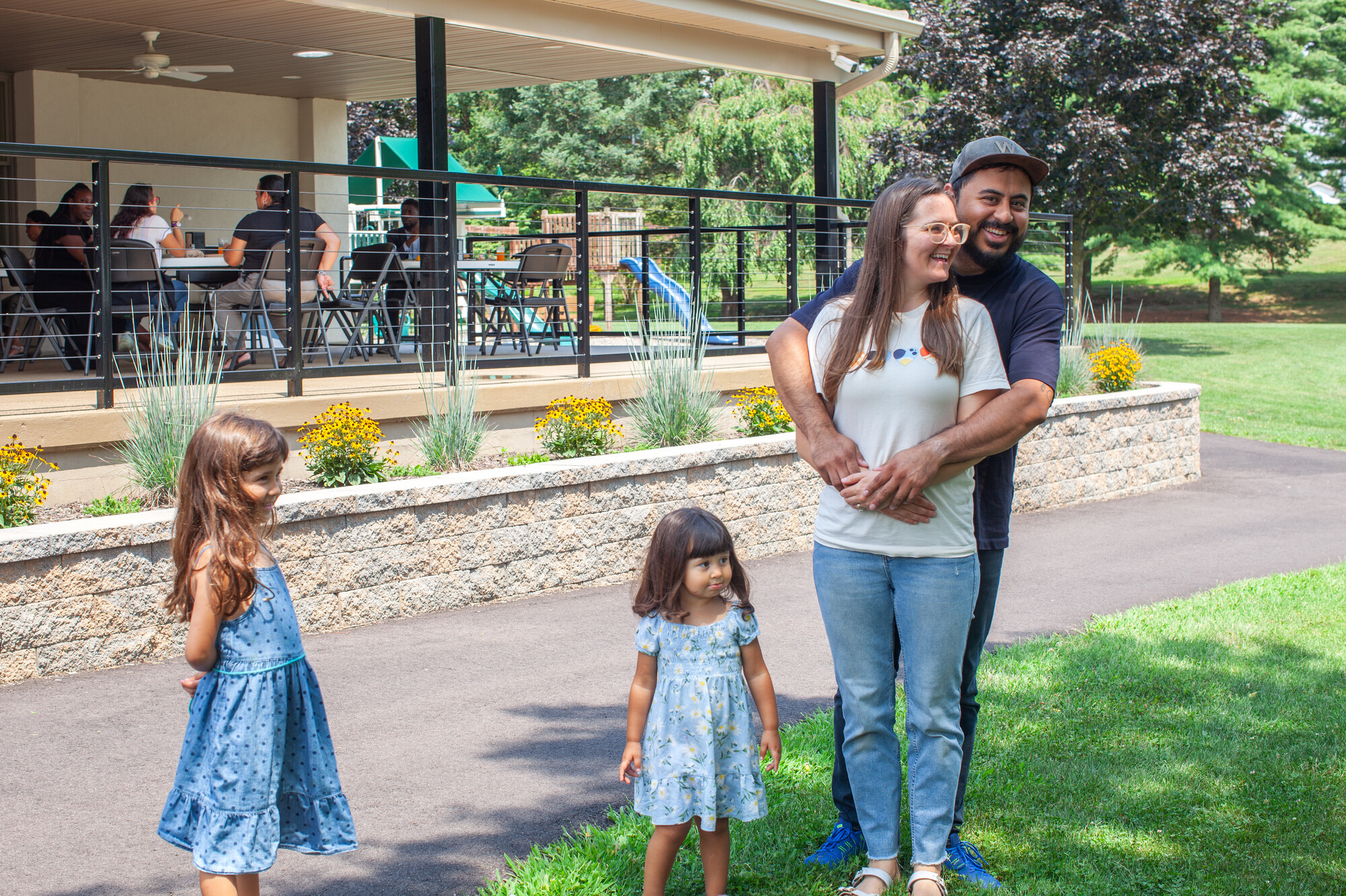 Two children and two adults stand outdoors in front of a building where people are eating. One adult has his arms around the other and they are both smiling.