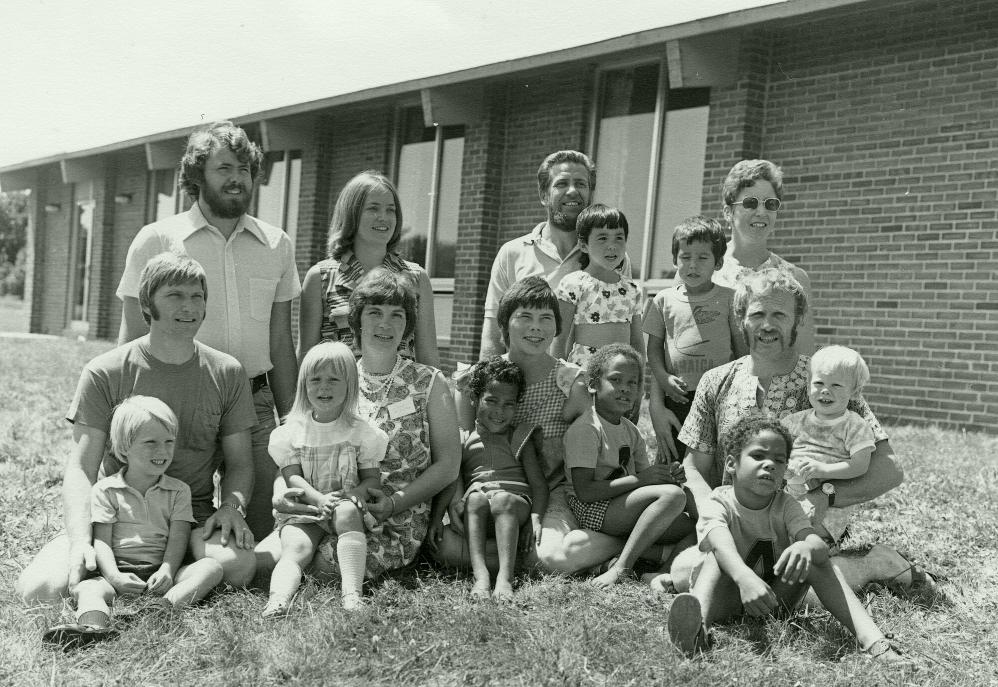 A group of adults and children poses outdoors in front of a brick building, arranged in two rows on grass.