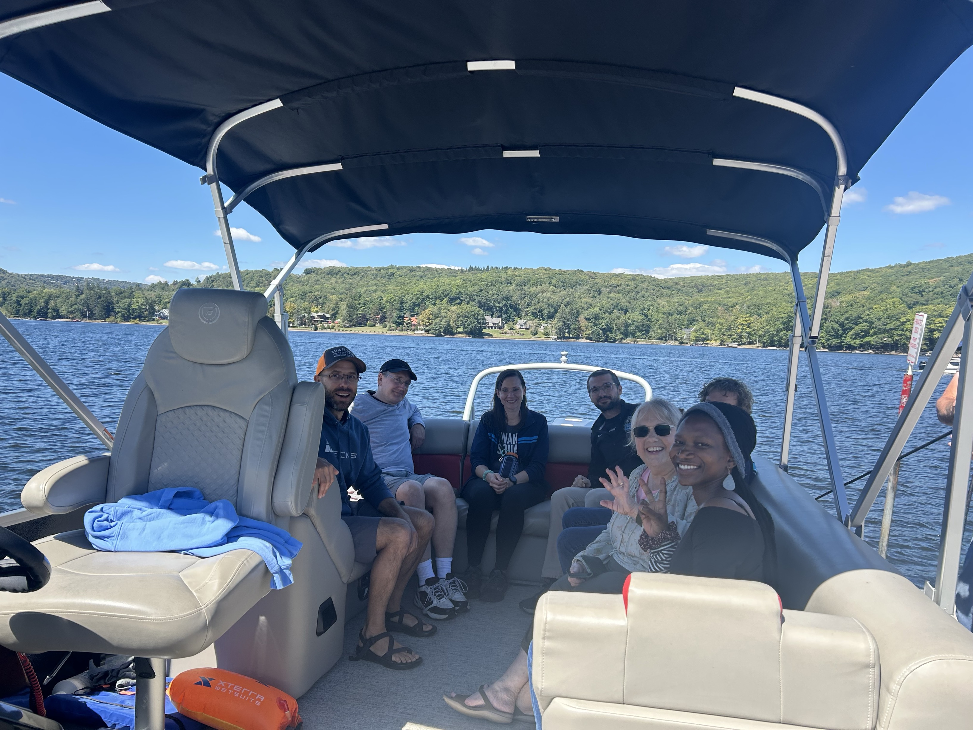 Seven people sit together on a boat in a like with green hills in the distance.