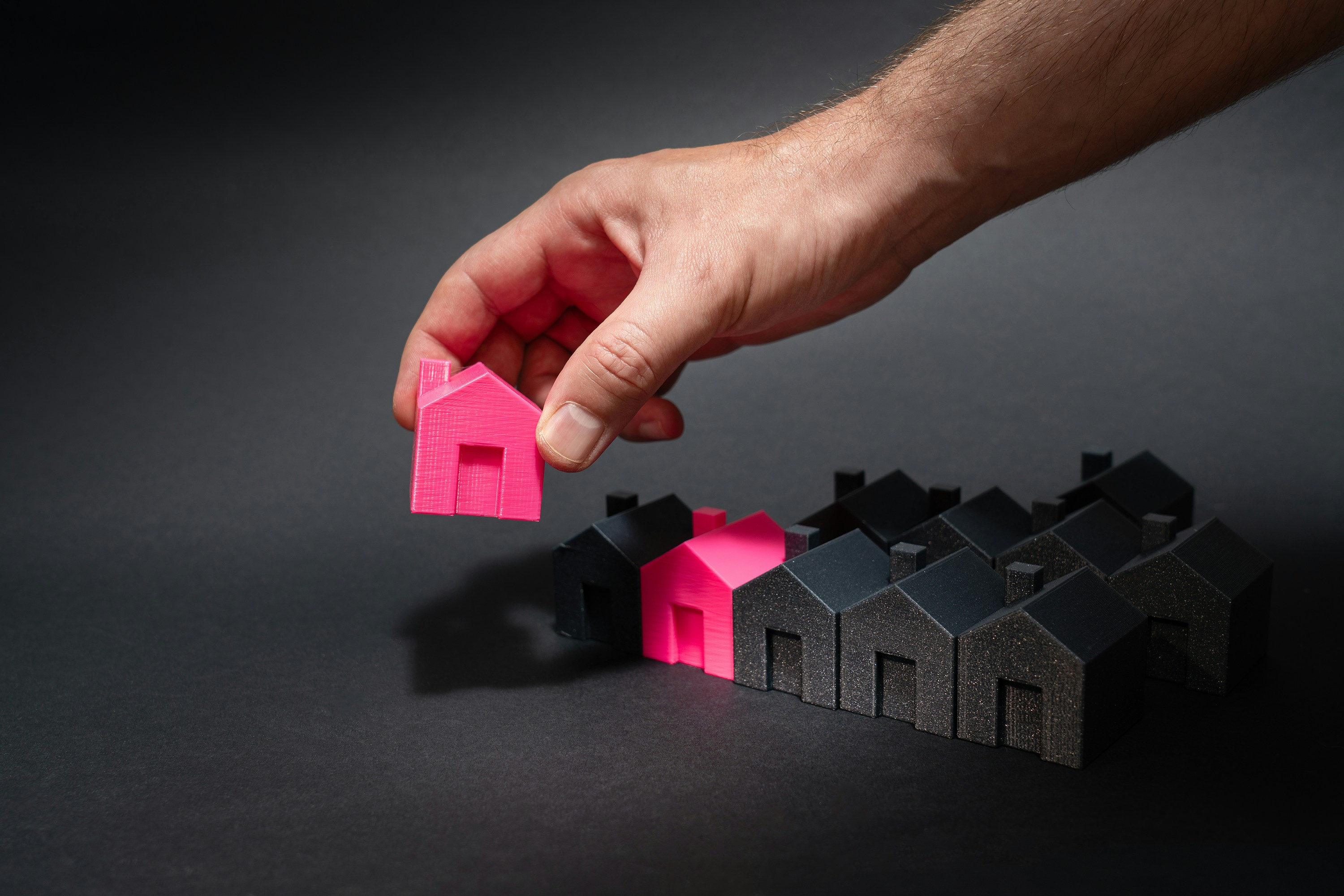 A hand holds up a pink model of a house from a row of black home models.