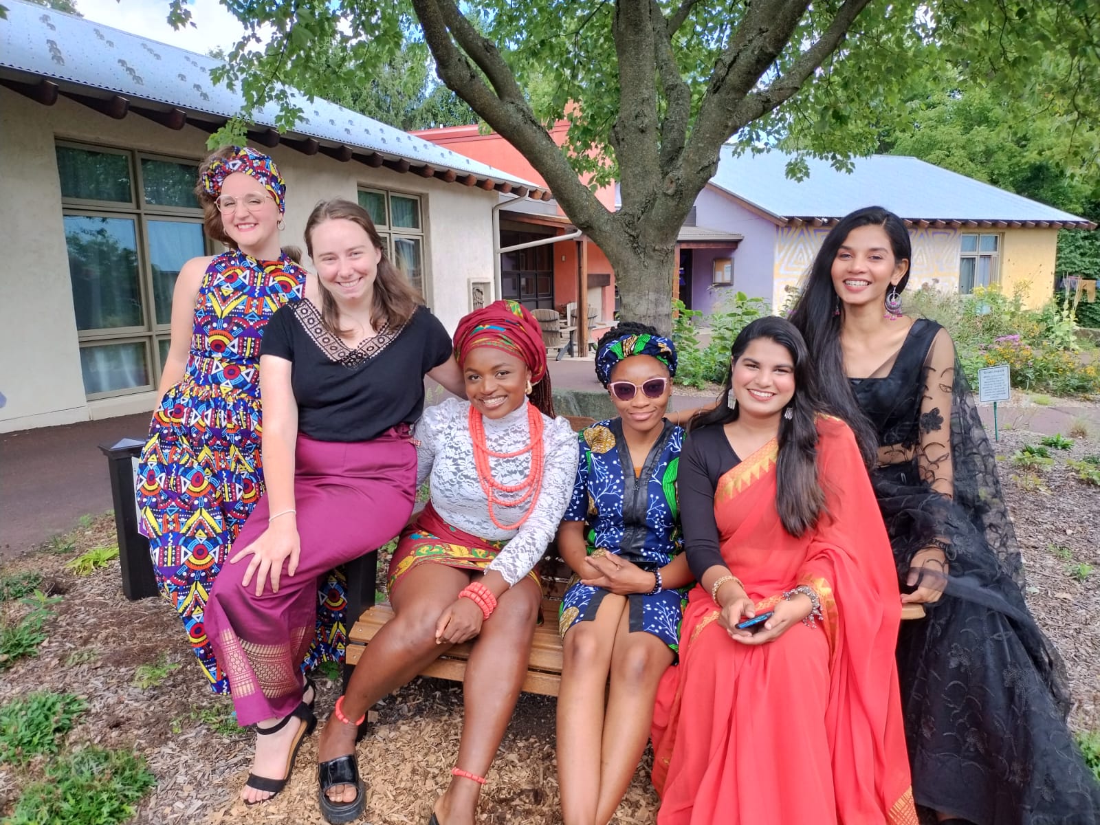 A group of five people wearing traditional clothing representing different cultures sit together in front of a tree.