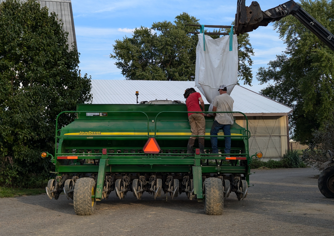 Two people stand on a large farm implement, filling it with seed.