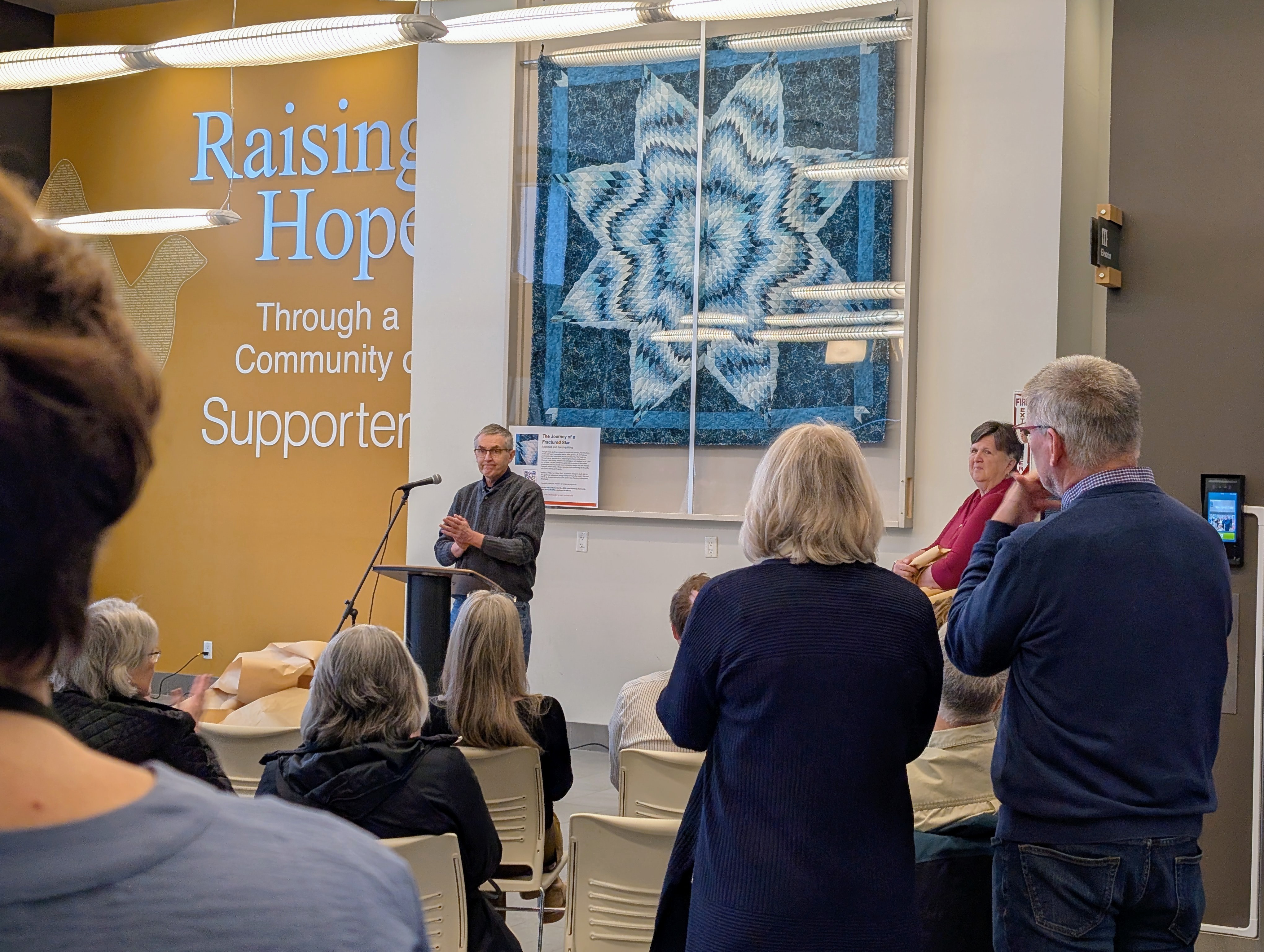 A crowd gathers in front of a man at a podium who is standing in front of a large quilt hanging on the wall.