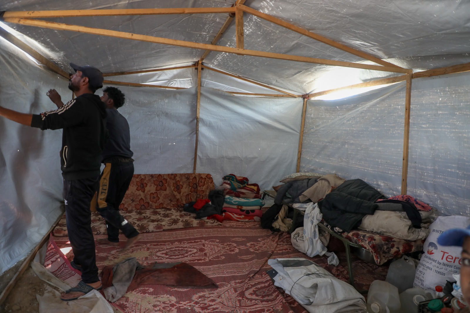 Two individuals adjust a tarp inside a tent. The space contains a patterned rug, bedding, and various items scattered around.