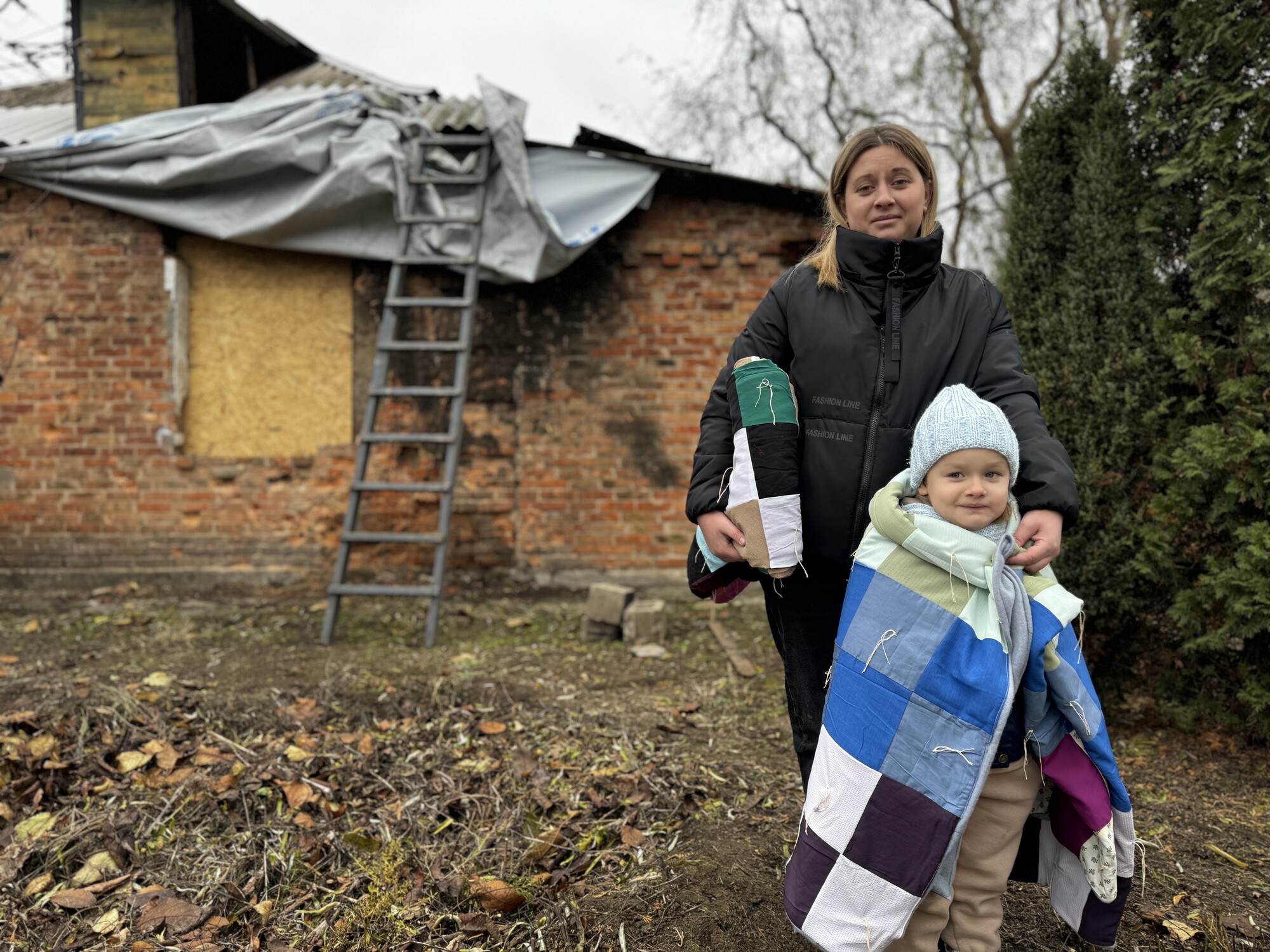 woman holding a comforter and 4-year-old daughter wrapped in comforter in front of house with no roof or windows