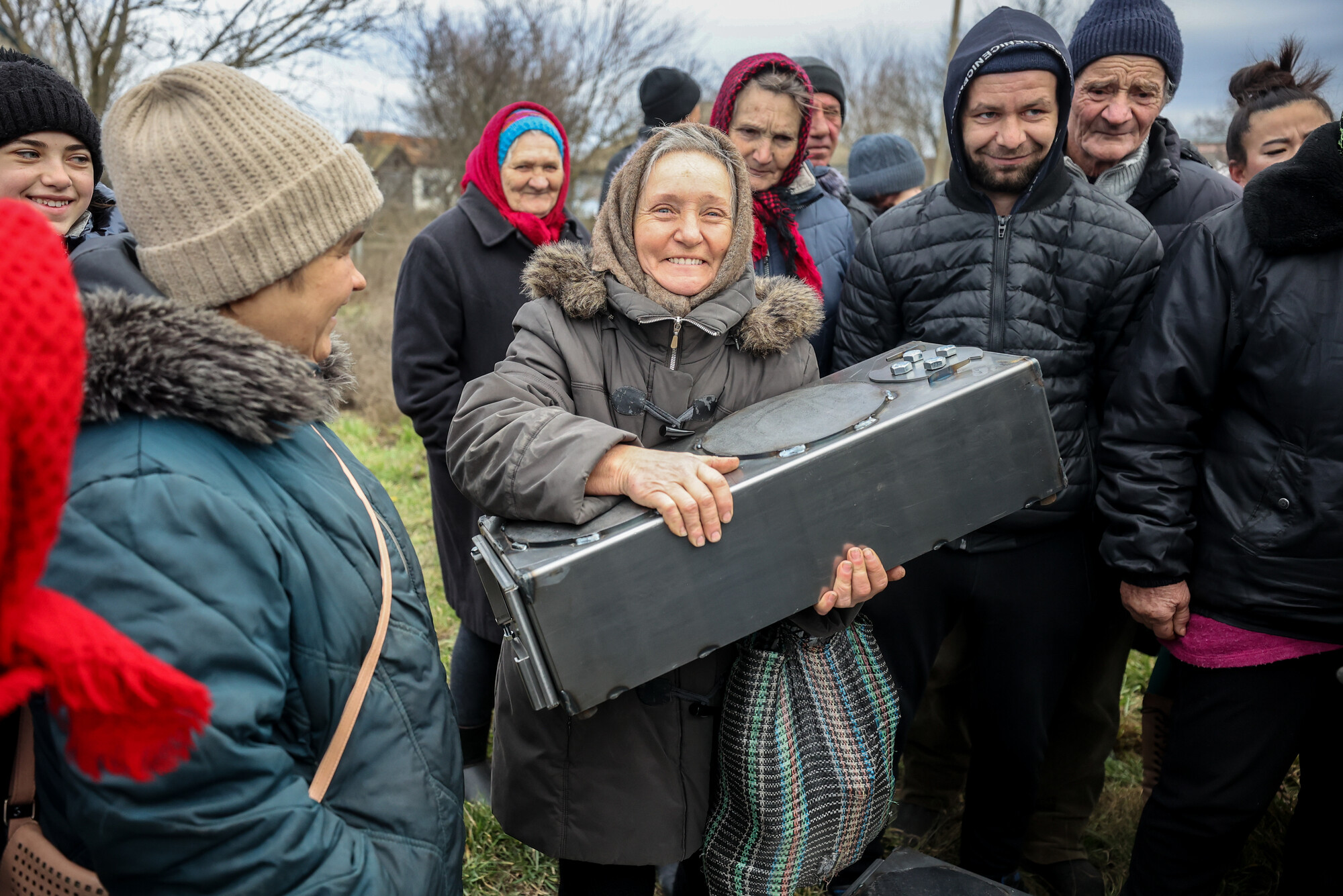 A woman in a village recovered from Russian military control in Ukraine smiles while holding a small wood stove.