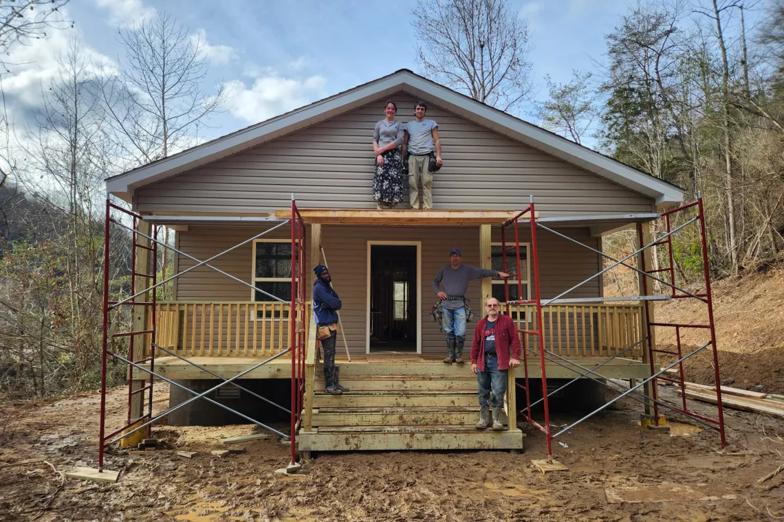 Five workers stand on the porch and roof of a house that is framed by scaffolding.