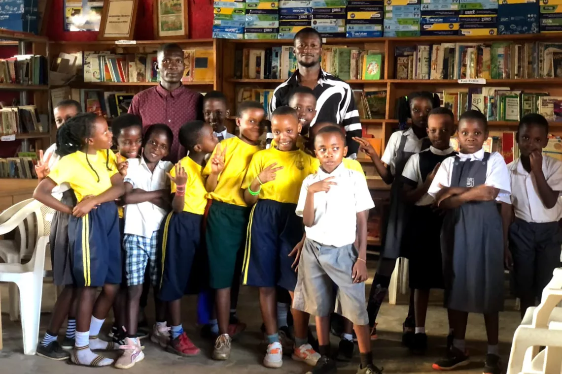 Two men stand behind a group of about 15 small children with bookshelves in the background.