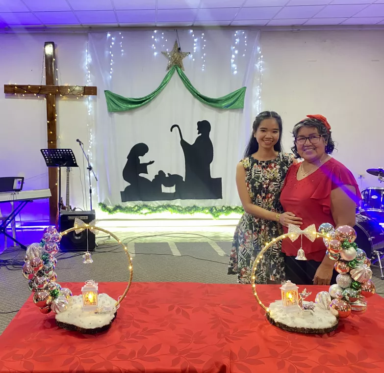 A mother and daughter stand behind a table in a church decorated for Christmas.