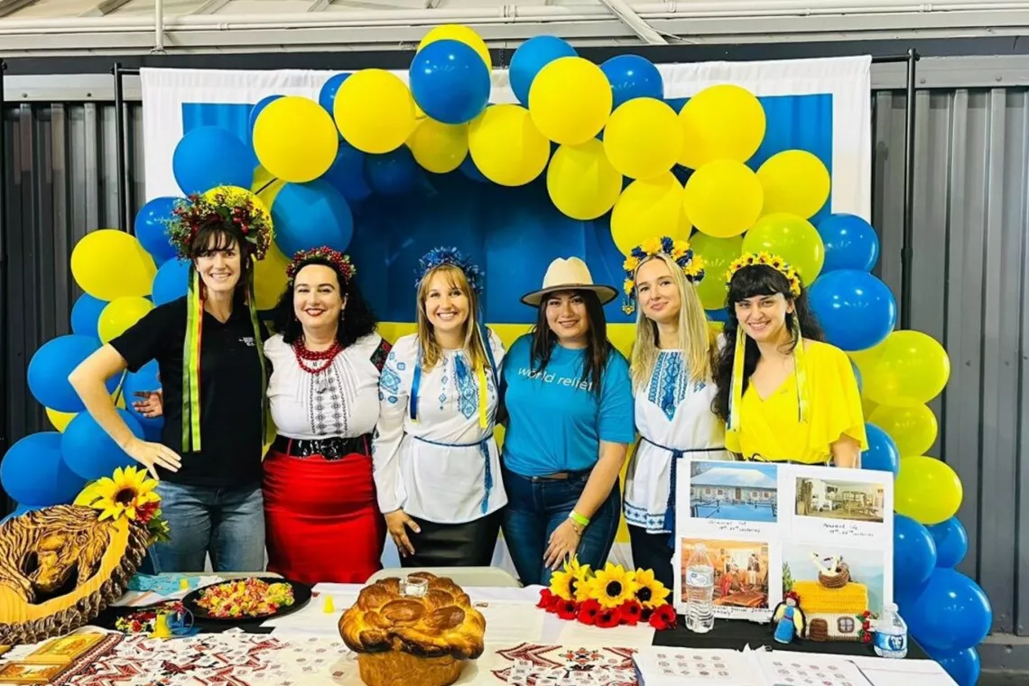 Six women wearing ethnic attire stand in front of an arc of blue and yellow balloons.