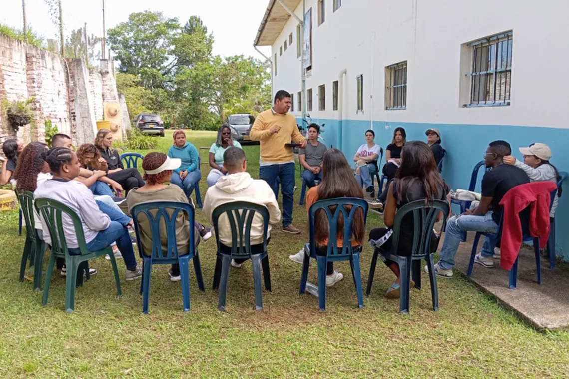 About 20 people sit in chairs in a circle with one person standing in the middle.