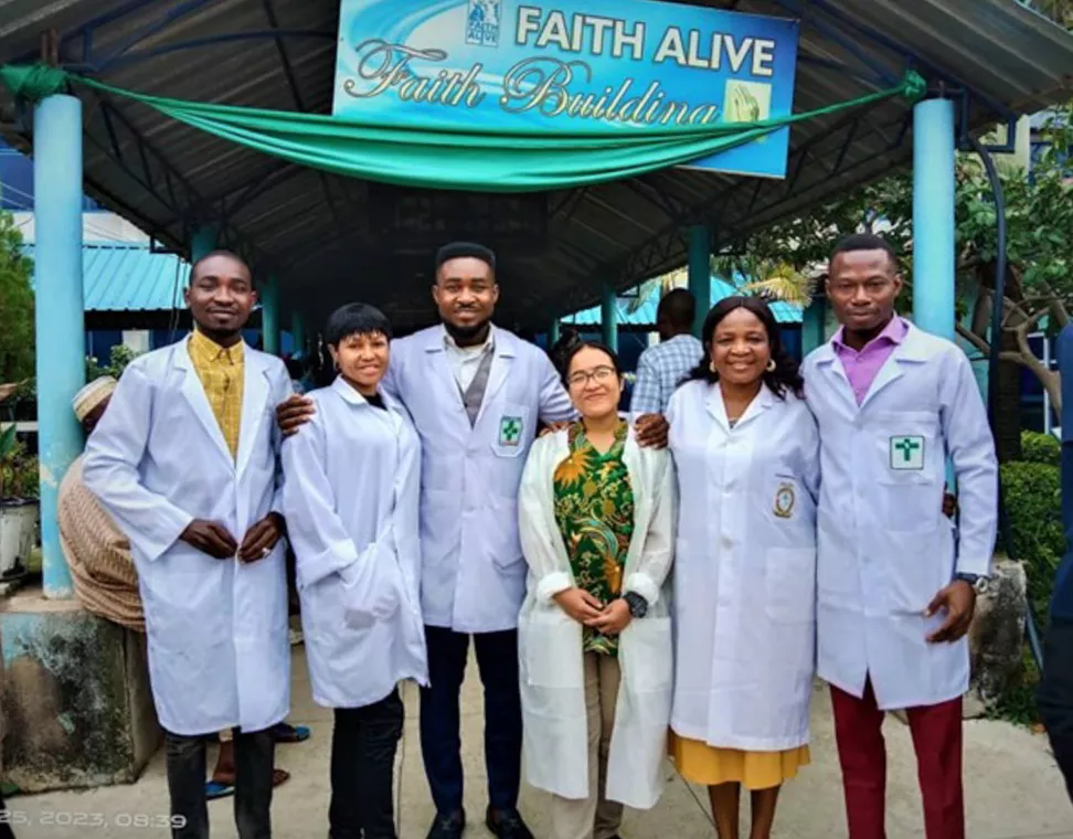 Six people in white medical coats stand outside beneath a sign that says, "Faith Alive Faith Building."