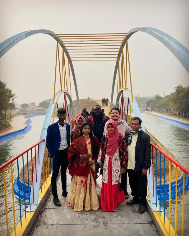 A group of people stands on a colorful bridge over a canal, with trees and a hazy sky in the background. They are dressed in a mix of traditional and casual attire.