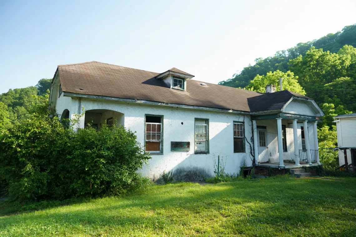 An old, neglected house with peeling white paint, surrounded by greenery, under a clear sky. It appears abandoned and in disrepair, with overgrown bushes.