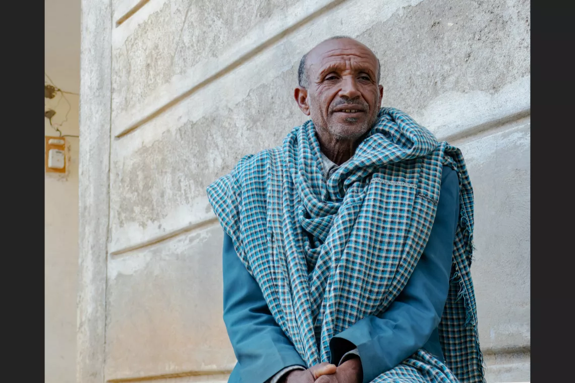 An elderly man wrapped in a checkered shawl looks pensively to the side against a pale wall under a clear sky.