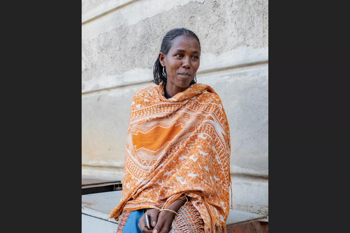 A woman sits outside, draped in an orange patterned shawl, looking to the side with a thoughtful expression. The background is a neutral-colored wall.