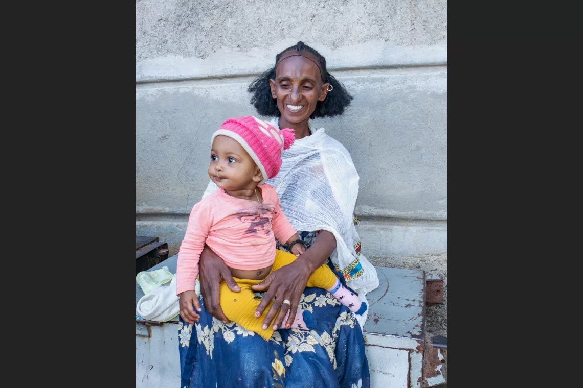 A woman in traditional attire smiles while holding a toddler wearing a pink hat. They are seated against a wall, suggesting a candid moment of familial warmth.