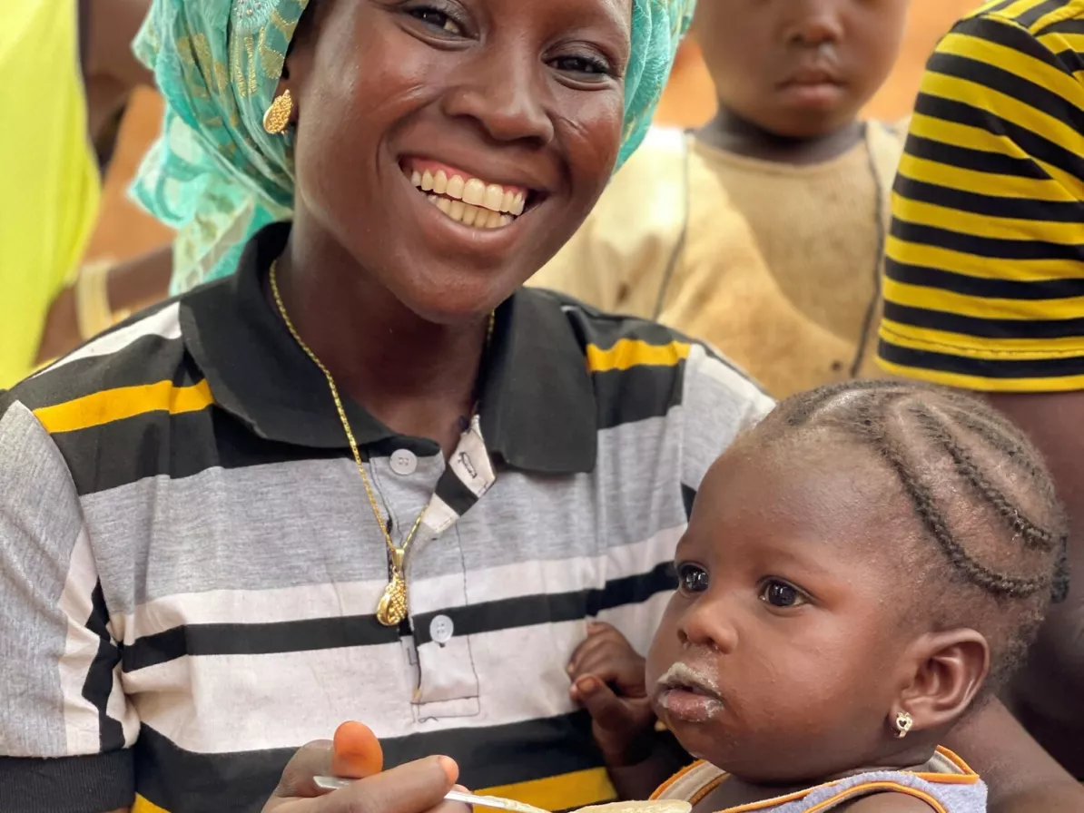 A woman sits and holds her baby. She is feeding her food from a yellow bowl and spoon.