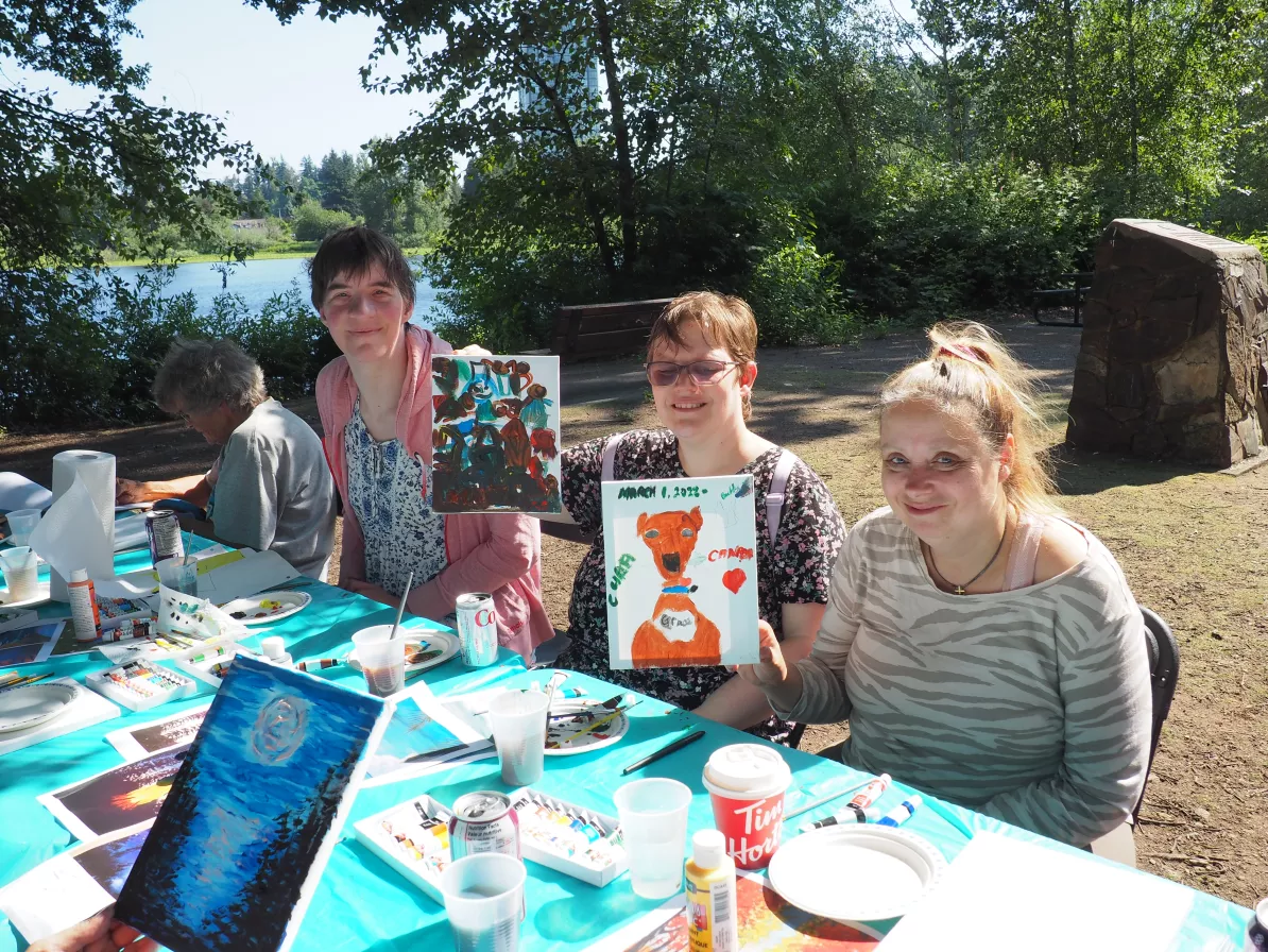 A group of three people painting at a table outside, one holds up their painting to show the camera.