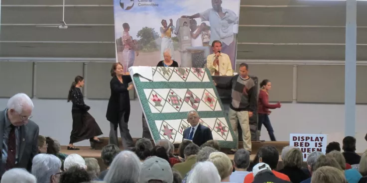 Three people display a quilt during an auction. Behind them is an auctioneer.