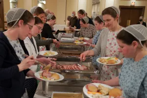 Ladies with traditional dress and head coverings gather around both sides of a buffet table.