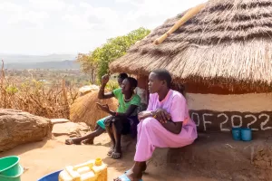 Three women sit in front of a hut home in Uganda.