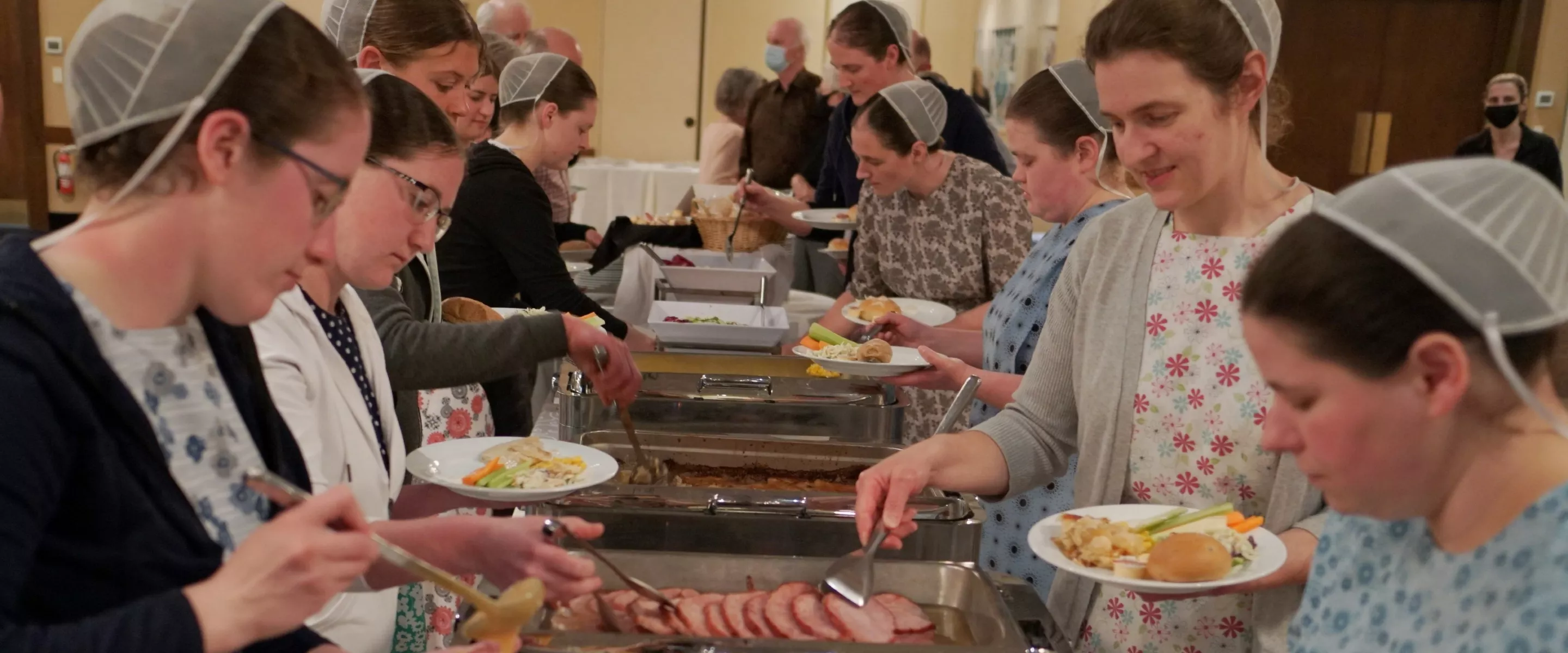 Ladies with traditional dress and head coverings gather around both sides of a buffet table.