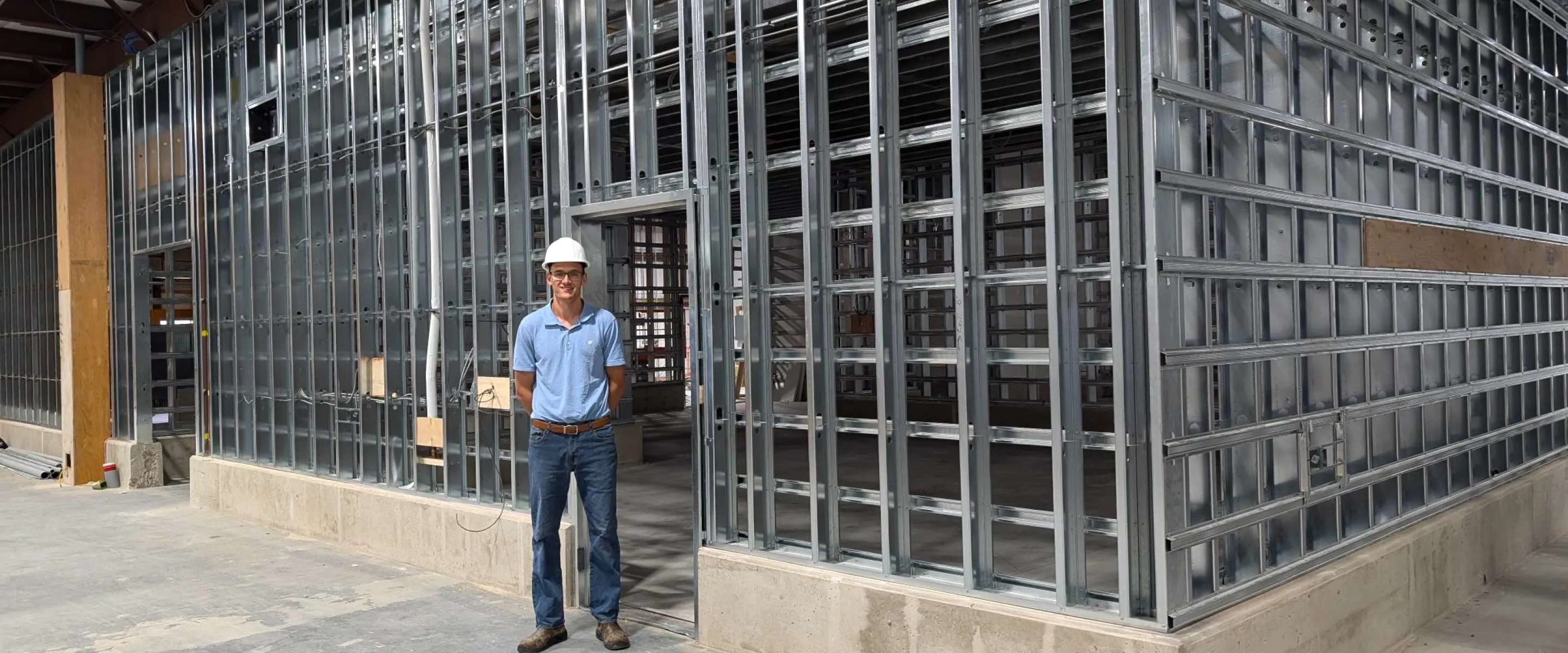 man in hard hat stands in front of a wall of steel studs under construction