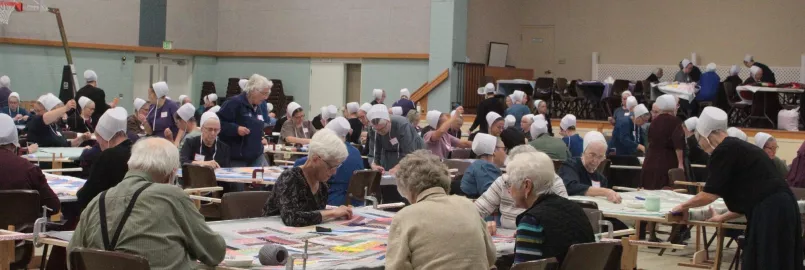 A large group of people sit around comforter tops and tie them.