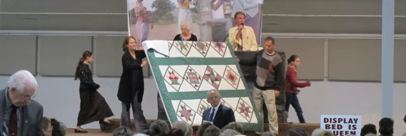 Three people display a quilt during an auction. Behind them is an auctioneer.