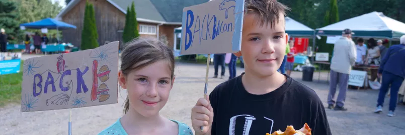 A boy and a girl hold up hand made cardboard signs that read Back Bacon, while the boy eats bacon on a bun.